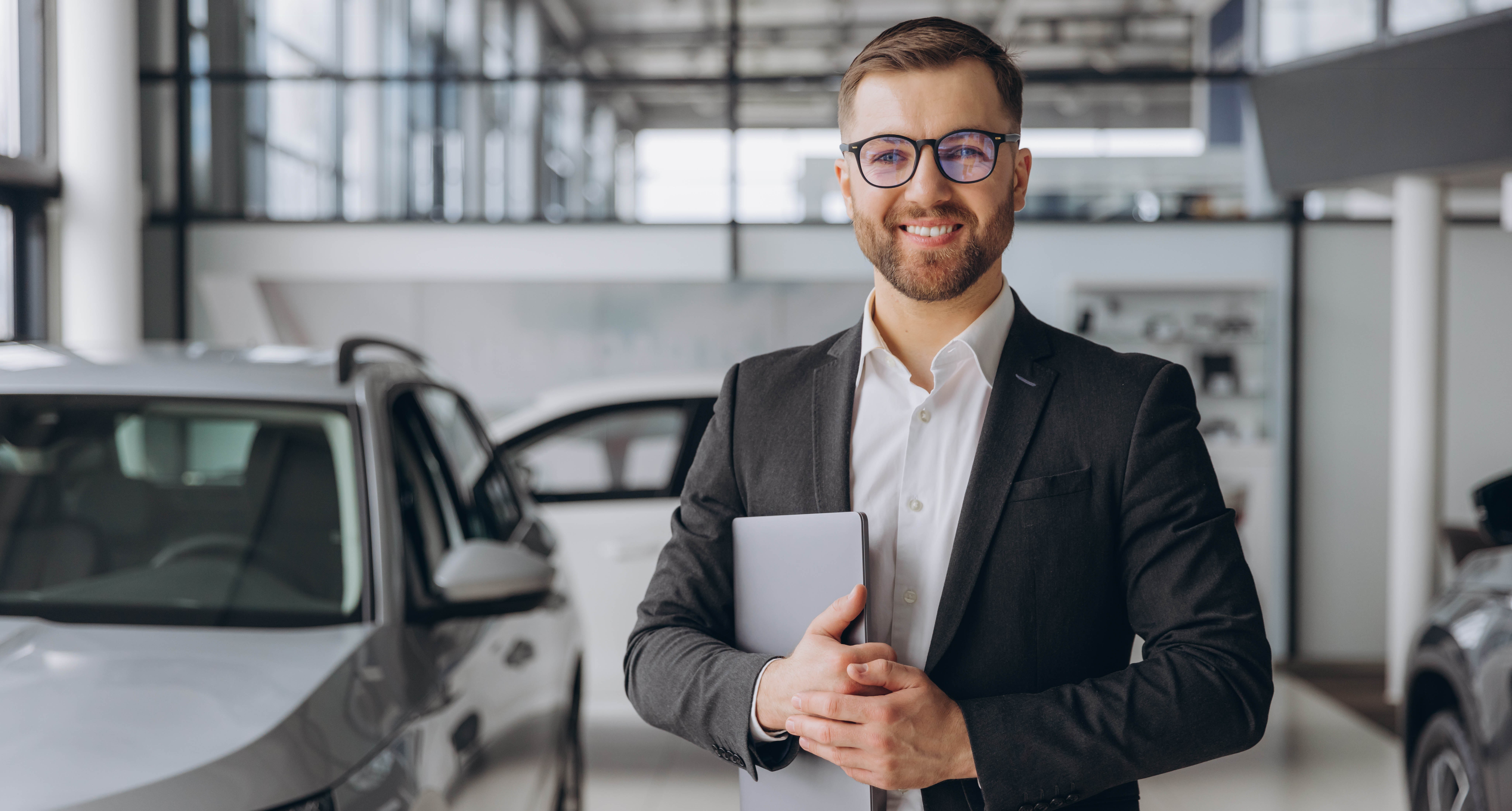 Modern bearded man glasses suit vehicle sales consultant using laptop inside car dealership copy